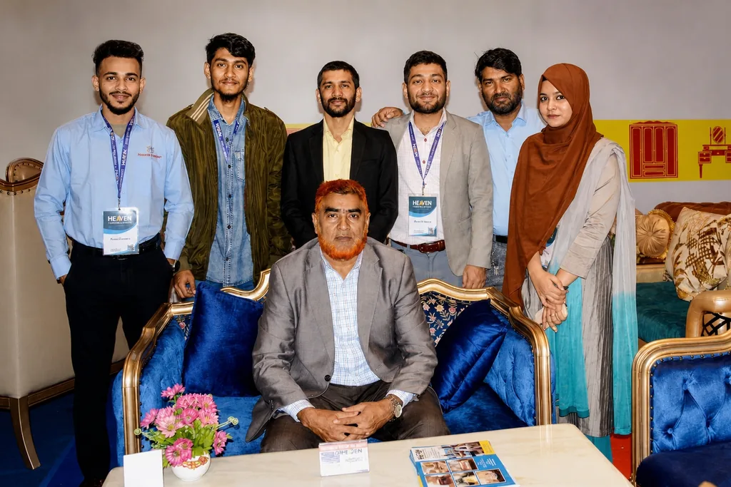 The Heaven Furniture Mart team at the International Furniture Fair in Chattogram: six staff standing behind Abul Kalam Bhuiyan, who is seated on a blue velvet carved settee